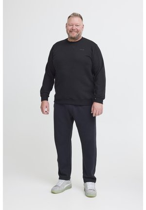 Un uomo sorride indossando un felpa nera, pantaloni neri e sneakers grigie chiare su uno sfondo bianco semplice.