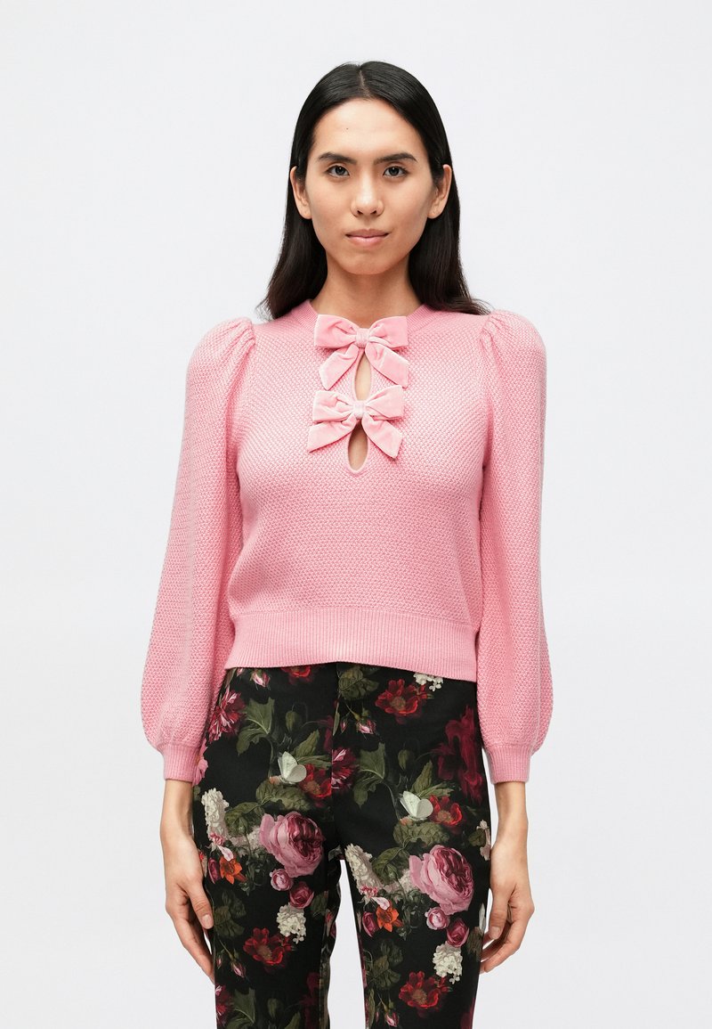 Rozā adīts džemperis ar pūkainām piedurknēm, ar diviem dekoratīviem lokiem un atvērumu priekšpusē, kopā ar biksēm ar ziedu rakstu.