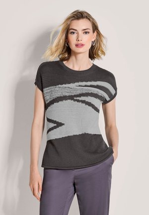 Vrouw met schouderlang blond haar draagt een zwart-grijs gebreid top met een abstract patroon en paarse broek, poserend met één hand in de zak.