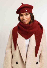 Beret de punto rojo con un pequeño moño y botón, combinado con una bufanda de lana roja. La bufanda presenta un diseño puntiagudo, llevada sobre un abrigo beige.