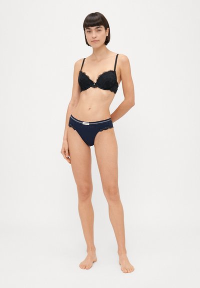 Reggiseno in pizzo nero con ferretto e bottoni decorativi, abbinato a mutandine blu navy con bordo in pizzo e dettaglio del logo sulla vita.