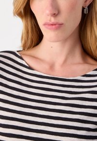 Fekete-fehér csíkos felső, széles nyakkivágással, puha anyagból készült. A modellen ezüst karika fülbevalók vannak, haján textúra és mozgás látható.
