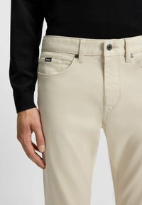 Beige cotton trousers featuring a slim fit, front pockets, and a black "BOSS" label on the left pocket with metal accents.