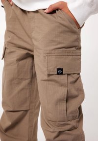 Beżowe spodnie bojówki wykonane z lekkiego materiału, z bocznymi kieszeniami i małą czarną naszywką z logo na kieszeni. Subtelna tekstura w kratkę na całej powierzchni.