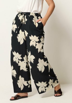 Osoba nosząca czarne spodnie z szerokimi nogawkami z dużym beżowym kwiatowym wzorem, biały top, brązowe klapki, stojąca z jedną ręką w kieszeni.