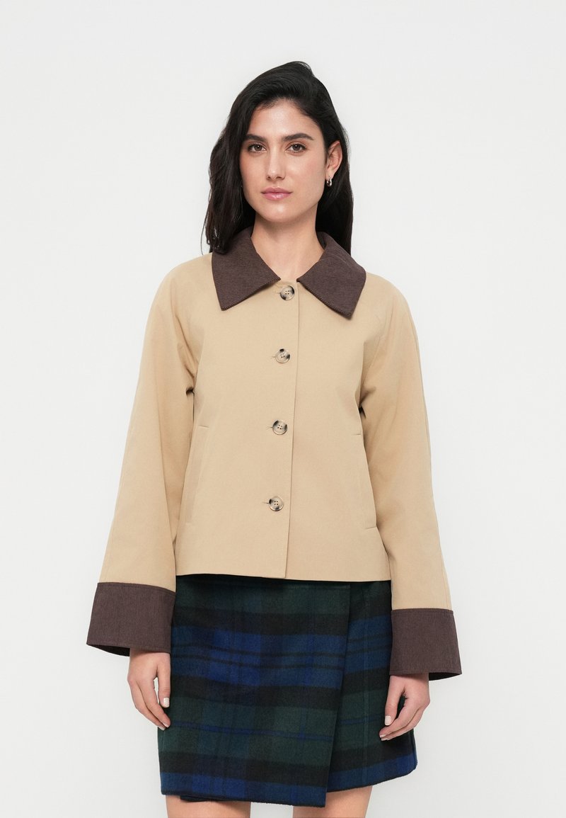 Μπεζ κοντό μπουφάν με γιακά και μανσέτες από σκουρό καφέ κορντόρι, με τέσσερα κουμπιά και πλαϊνές τσέπες. Συνδυάζεται με φούστα καρό.