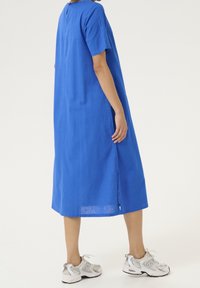 Sininen pellavamekko, jossa on lyhyet hihat, polvipituus, sivuhalkio ja pieni avonainen yksityiskohta takana, yhdistetty harmaisiin urheilukengiin.