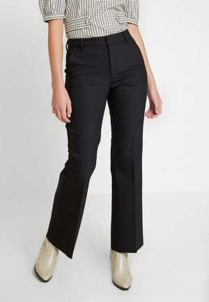 Mujer con pantalones negros de pierna recta hechos a medida, botines de cuero de serpiente beige y una blusa de manga corta a cuadros, de pie contra un fondo liso.