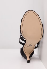 Sandale noire à talon haut reposant sur le côté, montrant une semelle beige lisse, un talon fin et plusieurs brides avec de petites décorations.