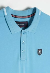 Błękitna koszulka polo z fakturowanej tkaniny, z trzema guzikami i małą naszywką z emblematem lwa po lewej stronie. Akcenty w postaci pasków na kołnierzyku.