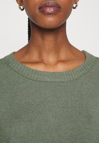 Grön stickad tröja med ribbad rund halsringning, som har en mjuk textur och jämn sömnad över hela plagget. Inga utmärkande dekorationer.