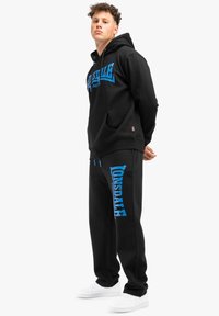 Črna kapuca s modrim logotipom spredaj, v kombinaciji z usklajenimi črnimi trenirkami, ki imajo modri dizajn. Bele superge dopolnjujejo obleko.