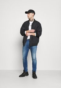 Black bomber jacket, white t-shirt with logo, blue jeans, black cap, and black sneakers. Casual outfit with varied textures and colors.