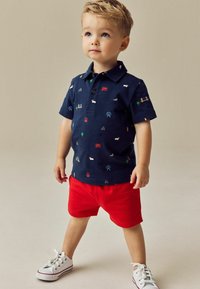Marineblå polo-shirt med forskellige farverige mønstre, parret med lyserøde shorts. Hvide sneakers fuldender outfittet, der viser en afslappet stil.