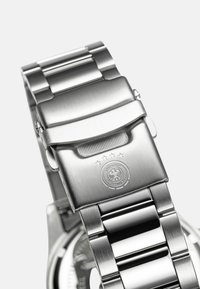 Silbernes Edelstahl-Armband mit gebürsteter Oberfläche, ausgestattet mit einer faltbaren Schließe, graviertem Logo und abwechselnd polierten Gliedern für zusätzliche Textur.