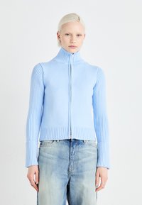 Svetlo modra jopica na zips z rebrasto teksturo, visokim ovratnikom in prilegajočimi rokavi, v kombinaciji s širokimi, svetlo pranimi jeans hlačami.