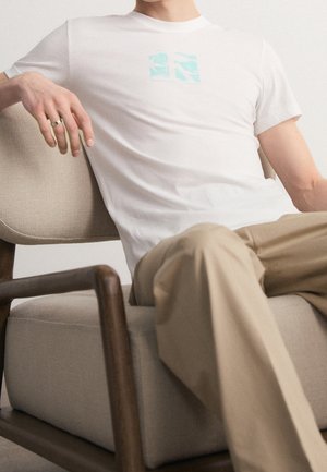 T-shirt blanc à manches courtes avec un graphique bleu clair sur la poitrine, associé à un pantalon beige, porté en position assise sur une chaise marron.