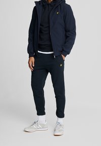 Marineblaue wasserdichte Jacke, schwarzer Kapuzenpullover und dunkle Jogginghosen mit gelbem Logo. Hellgraue Sneaker mit weißen Akzenten und graue Socken.