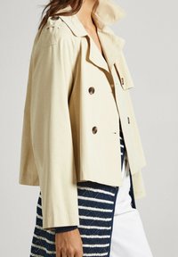 Abrigo trinchera recortado en color beige con diseño de doble botonadura, grandes botones y epaulets en los hombros. Tejido texturizado con un acabado suave.