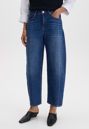Frau trägt hoch sitzende, gerade geschnittene blaue Denim-Jeans, schwarze Slip-on Pumps, weißes langärmeliges Hemd und schwarze Strickweste.