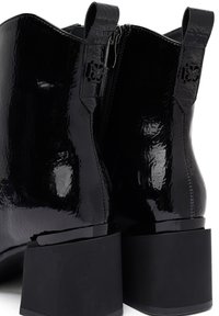 Botines de charol negro con punta cuadrada, cierre de cremallera y detalles en forma de lazo. Cuentan con un tacón grueso negro para mayor estabilidad.