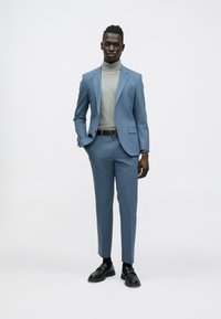 Világoskék öltöny szűk gallérral, szürke garbó és fekete öv. Fekete mokaszinok egészítik ki az öltözéket, amely összességében sima textúrájú.