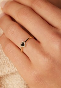 Bague en or avec une pierre noire en forme de cœur, entourée de petites pierres claires. Portée sur une main avec une surface texturée, sur un fond clair.