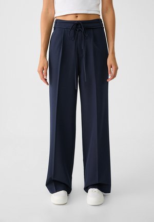 Persoon draagt marineblauwe wijde broek met voorplooien, taille met trekkoord, cropped wit topje en witte sneakers, staand op een grijze vloer.