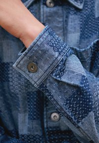 Mano infilata nel polsino di una giacca di jeans blu a pezzetti con motivi cuciti e bottoni a pressione in metallo.