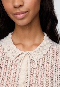 Hellbeige gestrickte Strickjacke mit einem gewellten Kragen, Bindemverschluss und einem filigranen Spitzenmuster mit Knopfdetails.