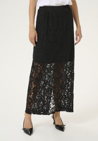 Jupe maxi en dentelle noire avec une taille élastique, présentant des motifs floraux complexes et un bas en tissu transparent. Portée avec des talons hauts noirs.
