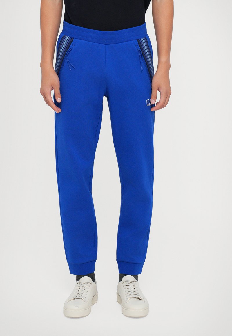 Blauwe sweatpants met een trekkoord in de taille, ritszakken en ribgebreide boorden. Gemaakt van zacht stof met verticale strepen langs de zakken.