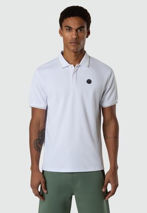 Mann trägt ein weißes kurzärmeliges Poloshirt mit einem kleinen runden Logo auf der Brust und grüne Hosen, steht vor einem grauen Hintergrund.