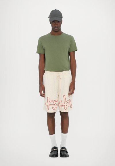 Green cotton t-shirt, beige shorts with red logo, white knee-high socks, and black loafers. Casual outfit suitable for warm weather.