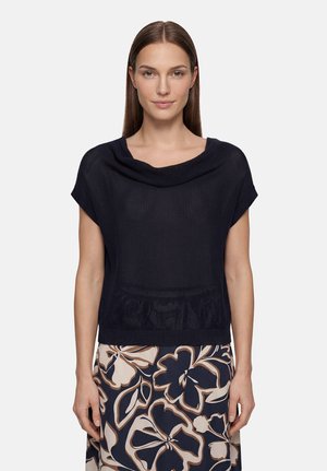 Femme aux cheveux bruns raides portant un haut en maille noire ample et une jupe à motifs avec de grands motifs floraux beige et bleu foncé.