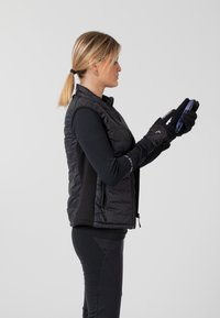 Schwarze Steppweste mit Reißverschluss, enganliegendes Langarmshirt und schwarze Handschuhe; hält ein Smartphone; Model mit Pferdeschwanz.