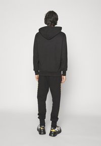 Černá mikina s kapucí na šňůrku, doplněná o odpovídající tepláky s bočními pruhy. Obuv tvoří robustní černé tenisky se vzorovaným designem.