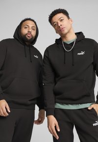 Sweatshirts à capuche noirs avec poches avant, ornés de logos Puma blancs. Fabriqués en matériau doux, portés avec un pantalon assorti.
