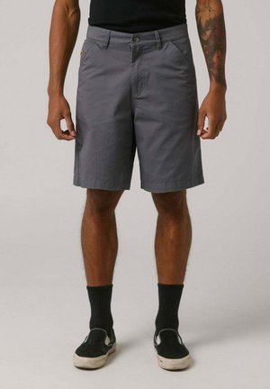 Uomo che indossa pantaloncini grigi a metà coscia, calzini neri e scarpe slip-on nere, in piedi con le braccia rilassate lungo i fianchi su uno sfondo semplice.