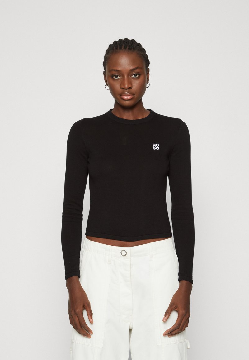 Černý dlouhý rukáv, ribby crop top s kulatým výstřihem a malým bílým logem na levé straně, spárovaný s bílými cargo kalhotami.