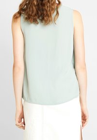 Haut sans manches vert menthe clair en tissu lisse, avec un ourlet arrondi et une coupe décontractée. Associé à une jupe de couleur crème.