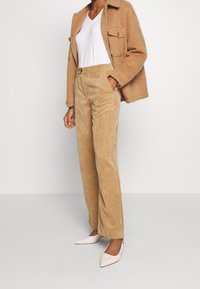 Pantalones de pana color beige de cintura alta combinados con una chaqueta de lana marrón y una camiseta blanca de corte en V. Los zapatos son tacones blancos de punta.