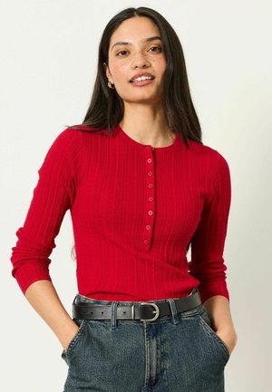 Kvinde med langt mørkt hår iført en rød knappet sweater og blå jeans med sort bælte, stående med hænderne i lommerne.