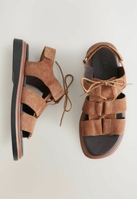 Sandalias de ante marrón con múltiples correas, un diseño de encaje y una suela marrón oscuro. Presenta una forma plana y ancha con acentos minimalistas.