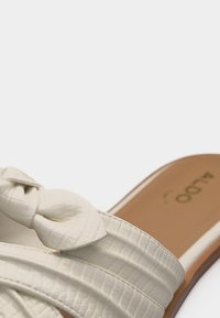 Gros plan sur une sandale slide blanche texturée avec une sangle nouée et une semelle intérieure beige affichant le nom de la marque ALDO.