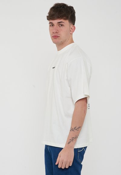 T-shirt en coton blanc surdimensionné avec des manches courtes et un col rond. Présente un petit texte noir sur la poitrine et un graphique sur la manche.