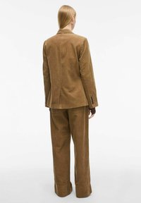Bruin corduroy pak met een op maat gemaakt colbert met notch-lapels en bijpassende broeken met een brede pijp. Geweven stof met verticale ribben.