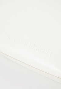 Bijela kožna torba s utisnutim logom "Calvin Klein" i suptilnim detaljima šivanja. Minimalistički dizajn, glatka tekstura.