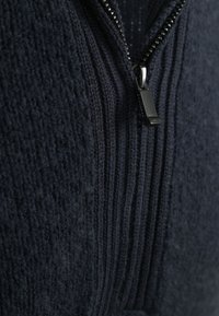 Close-up van een marineblauw gebreid kledingstuk met een gedeeltelijk gesloten zwarte metalen ritssluiting, waarbij de gestructureerde geribbelde en pluizige gebreide stof zichtbaar is.