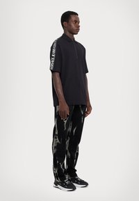 Čierna polokošeľa so zipsom a krátkymi rukávmi, na ramene s bielym nápisom "VERSACE JEANS COUTURE". V kombinácii s čiernymi džínami v štýle tie-dye a teniskami.